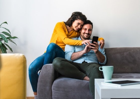A couple snuggling on a sofa thinking about the future while using a mobile phone to apply for a First-Time Homebuyer Loan.