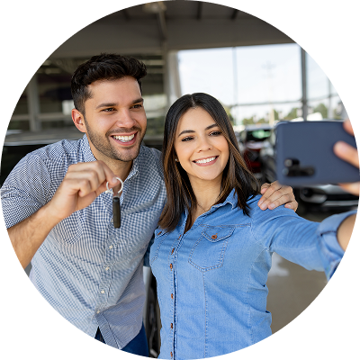 A smiling couple taking a selfie in front of their new car at a dealership, celebrating their first car purchase.