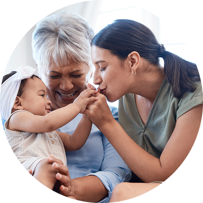 A mother kisses her baby’s hand while sitting on her grandmother’s lap, symbolizing generations—reminder to update your account beneficiaries.